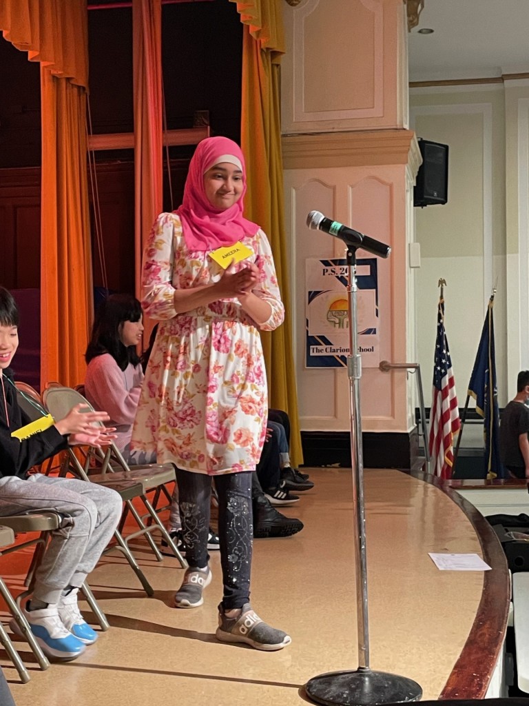 A student wearing a pink hijab and a floral tunic approaches the microphone