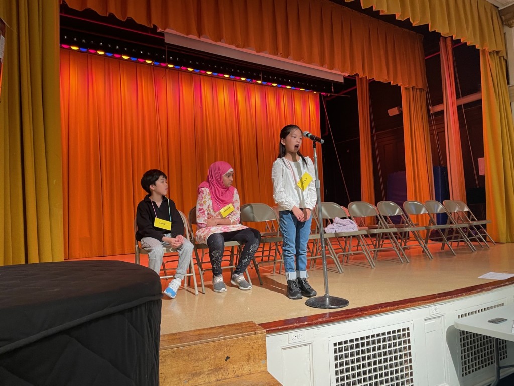 Two students sit on stage while a third student wearing a white shirt stands at the microphone