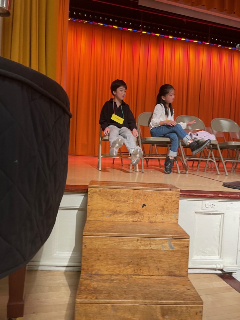 two students sit on stage. One is wearing a black shirt and one is wearing a white shirt