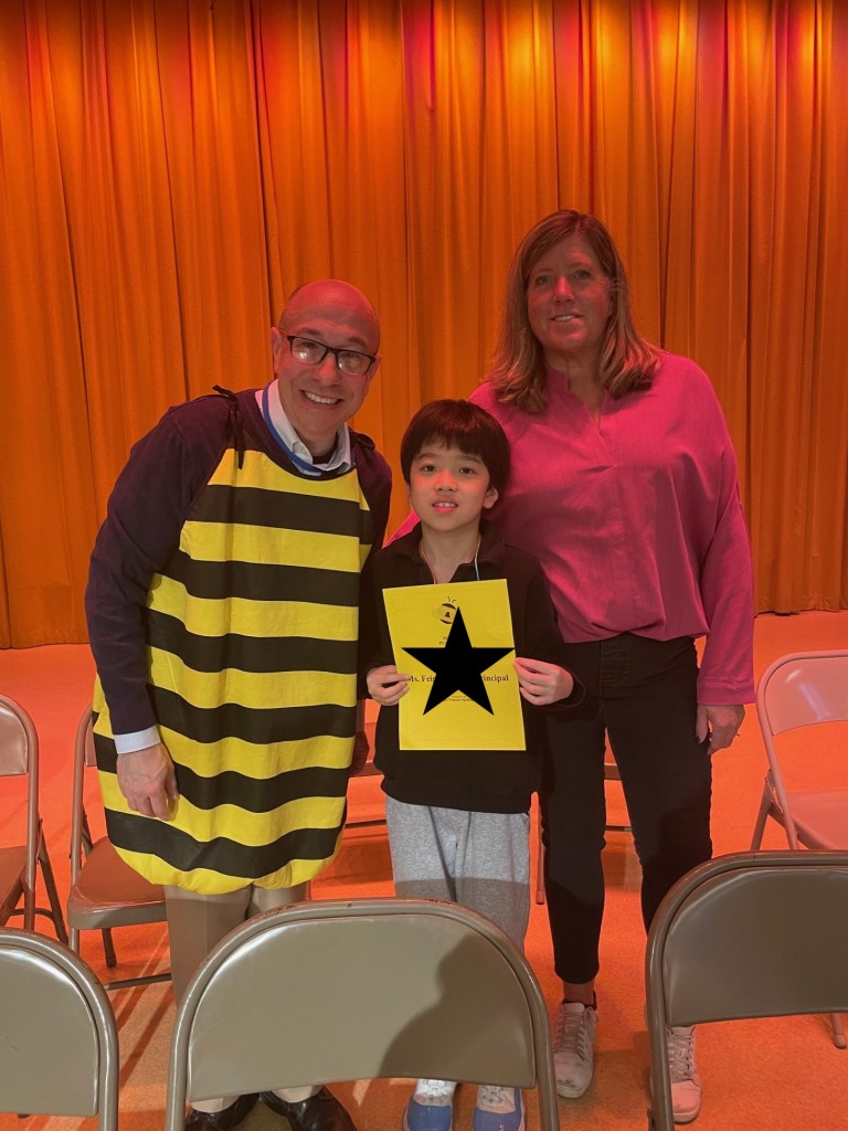 A man in a bumblebee costume stands with the winner of the spelling bee wearing a black shirt and a woman in a pink shirt